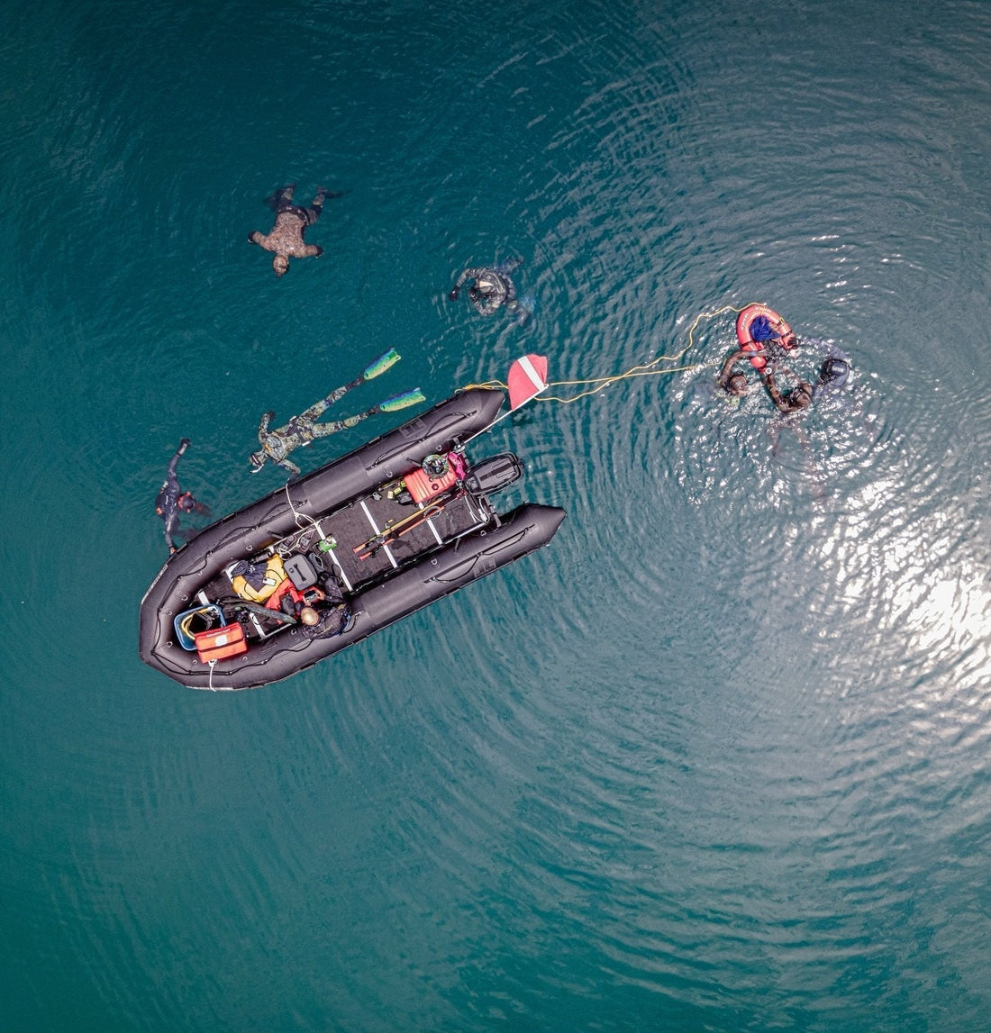 Aerial view of divers in camouflage wetsuits swimming near a black inflatable boat in clear water