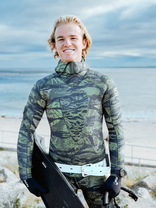 Smiling man in camouflage wetsuit holding spearfishing equipment by the beach with cloudy sky