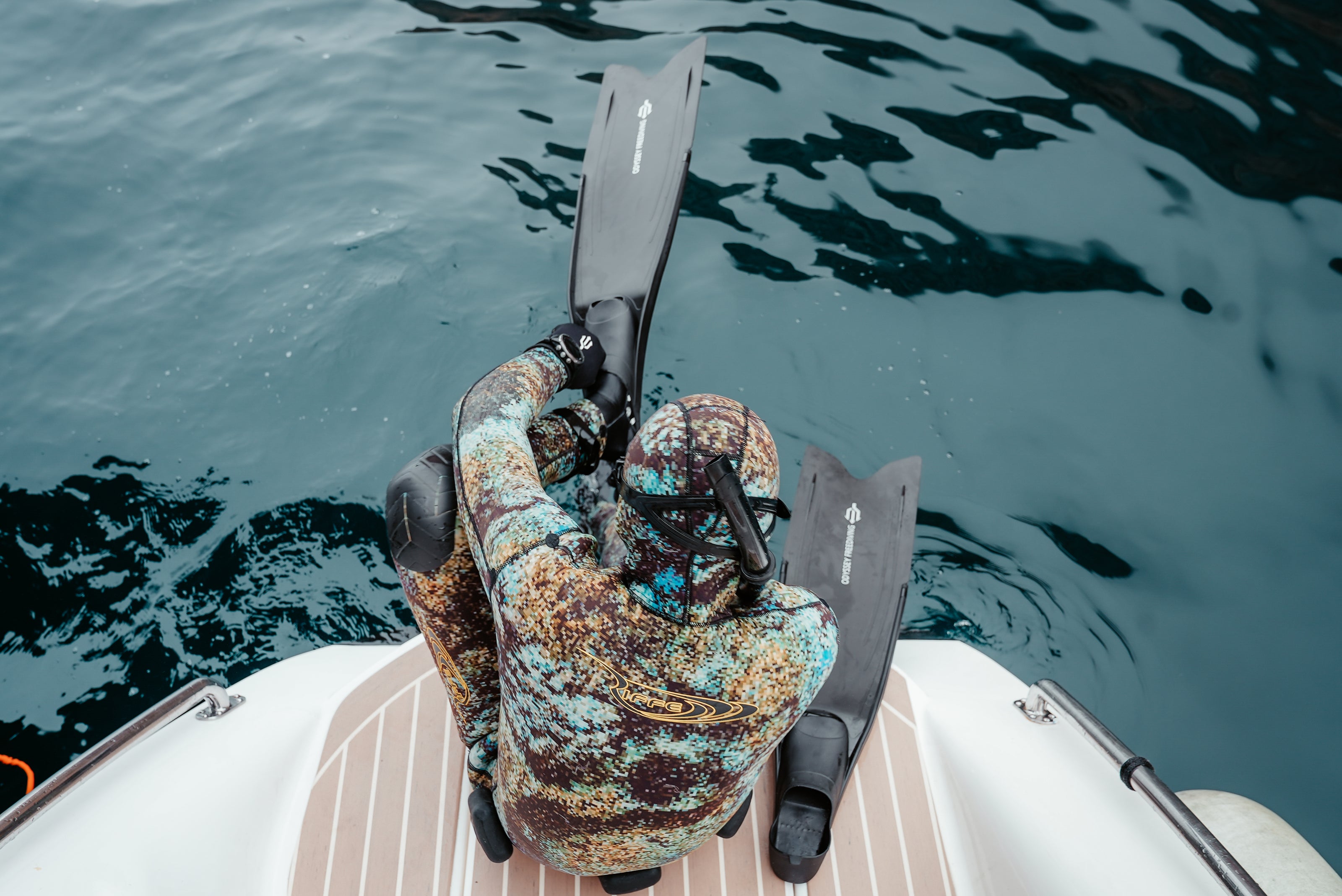 Person wearing camouflage wetsuit and black diving fins sitting on boat deck preparing to dive into water